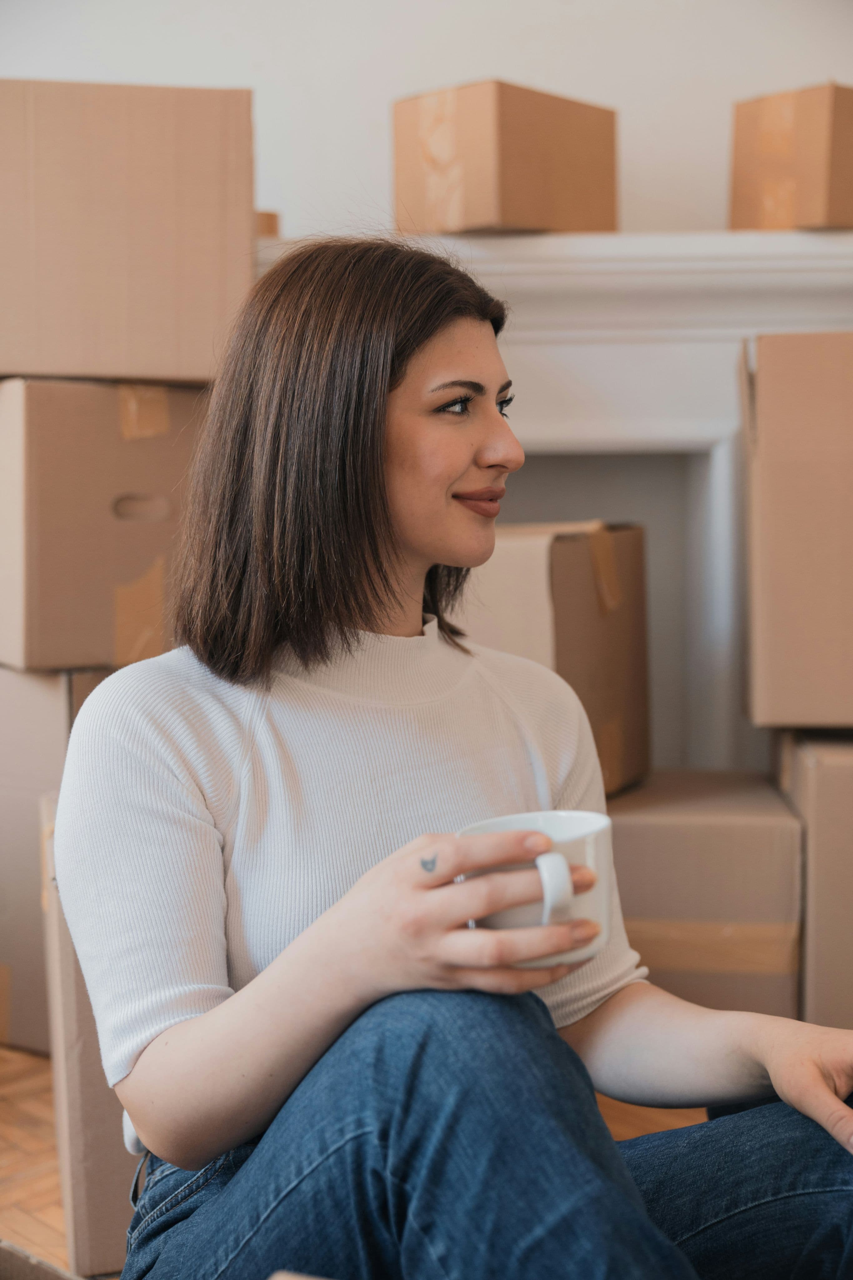 Femme souriante tenant une tasse blanche, assise devant des cartons de déménagement empilés.