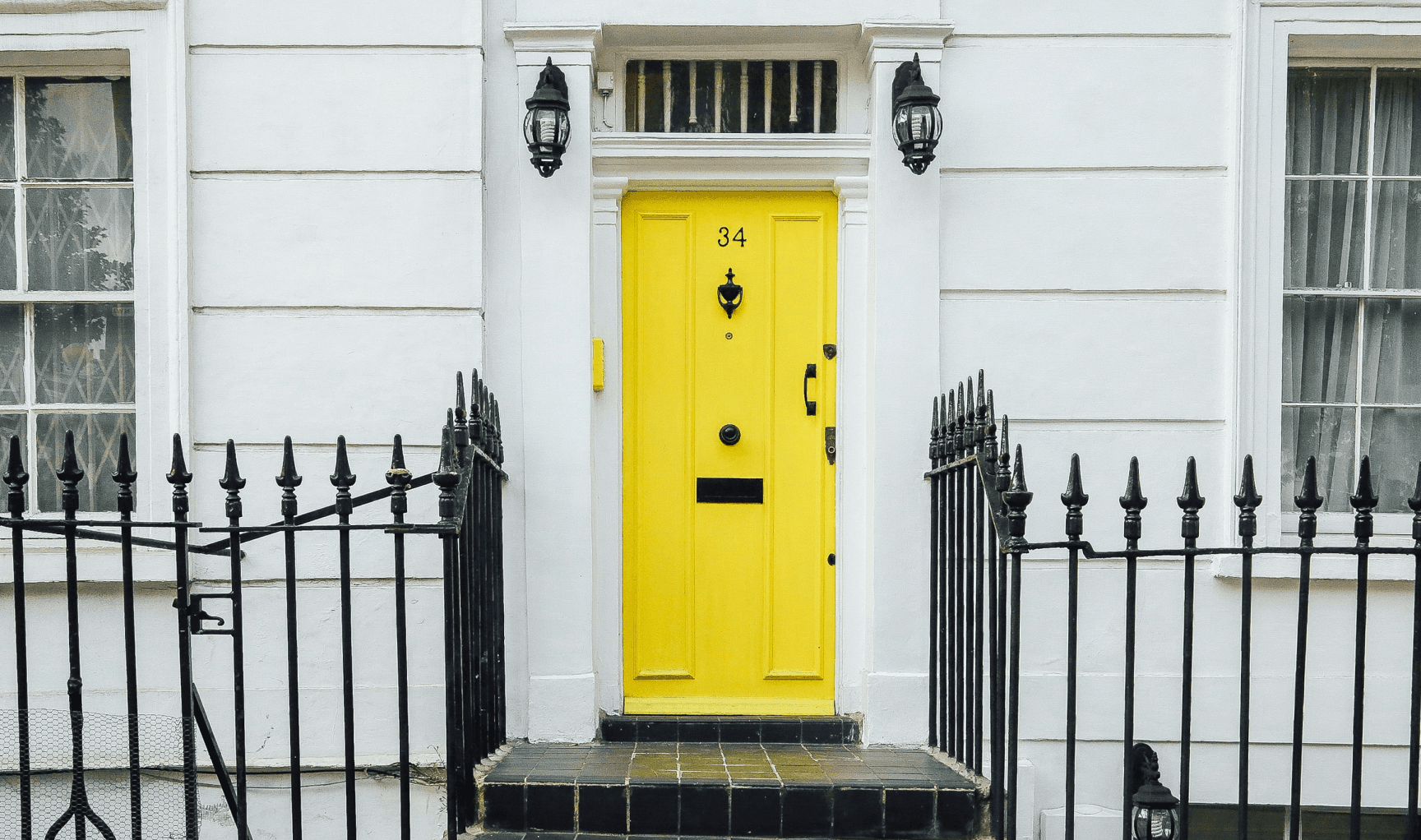 Porte d'entrée jaune vif avec le numéro 34, entourée d'une façade blanche classique et d'une clôture en fer forgé noir.