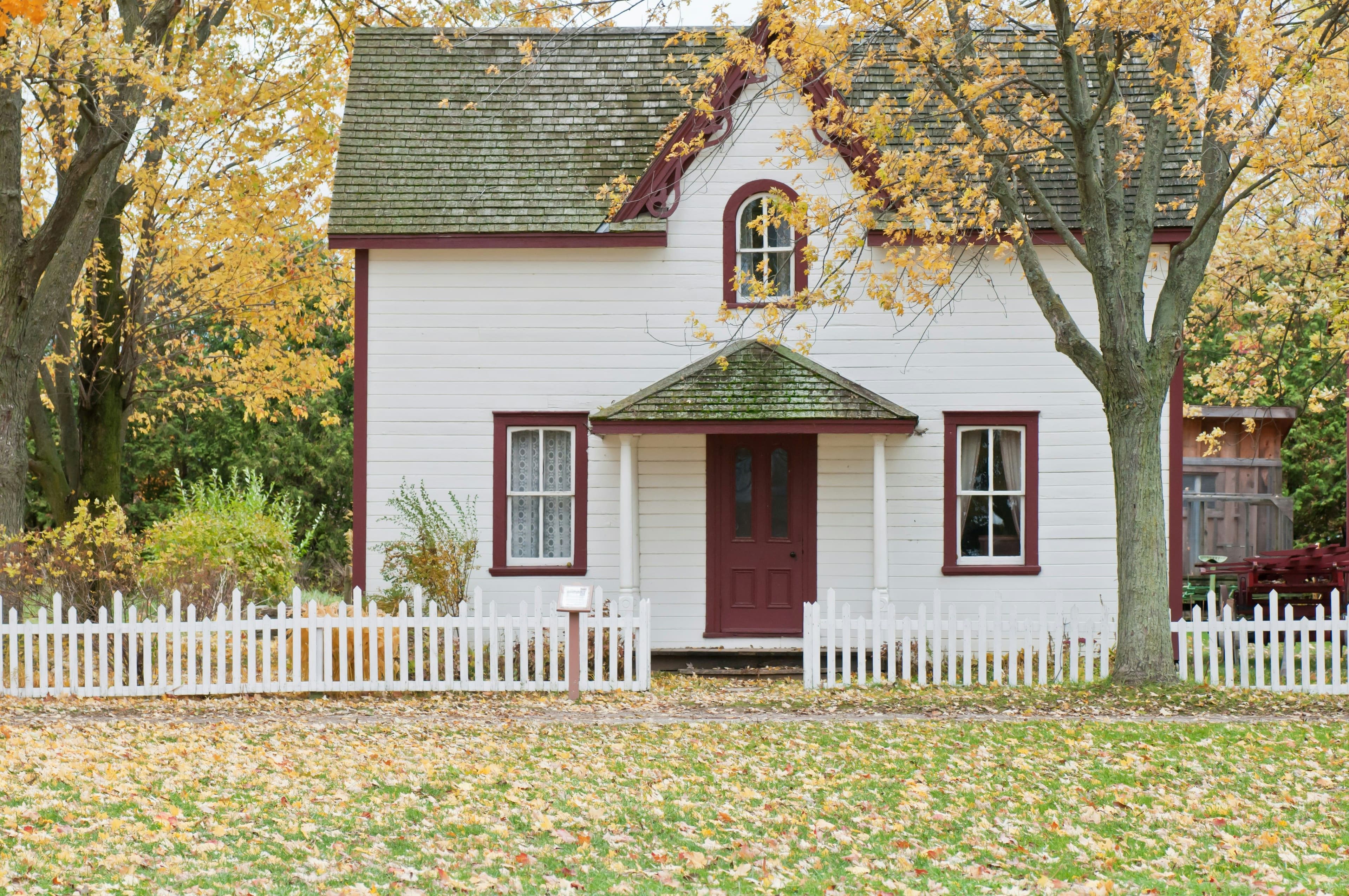 Maison blanche avec des encadrements rouges, entourée d'une clôture blanche, au milieu des arbres aux feuilles jaunes en automne.