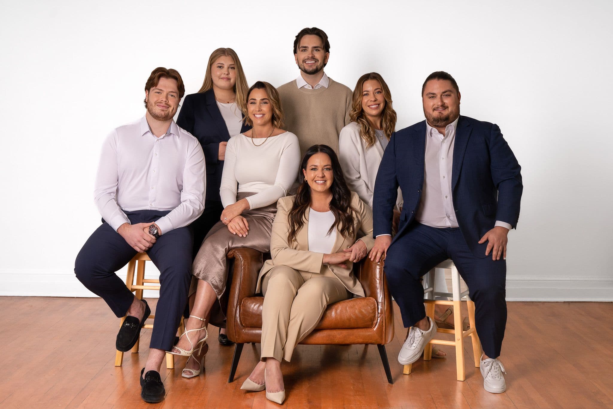 Groupe de sept personnes souriantes posant dans un studio, habillées de tenues professionnelles dans des tons neutres, sur des chaises et un fauteuil en cuir.