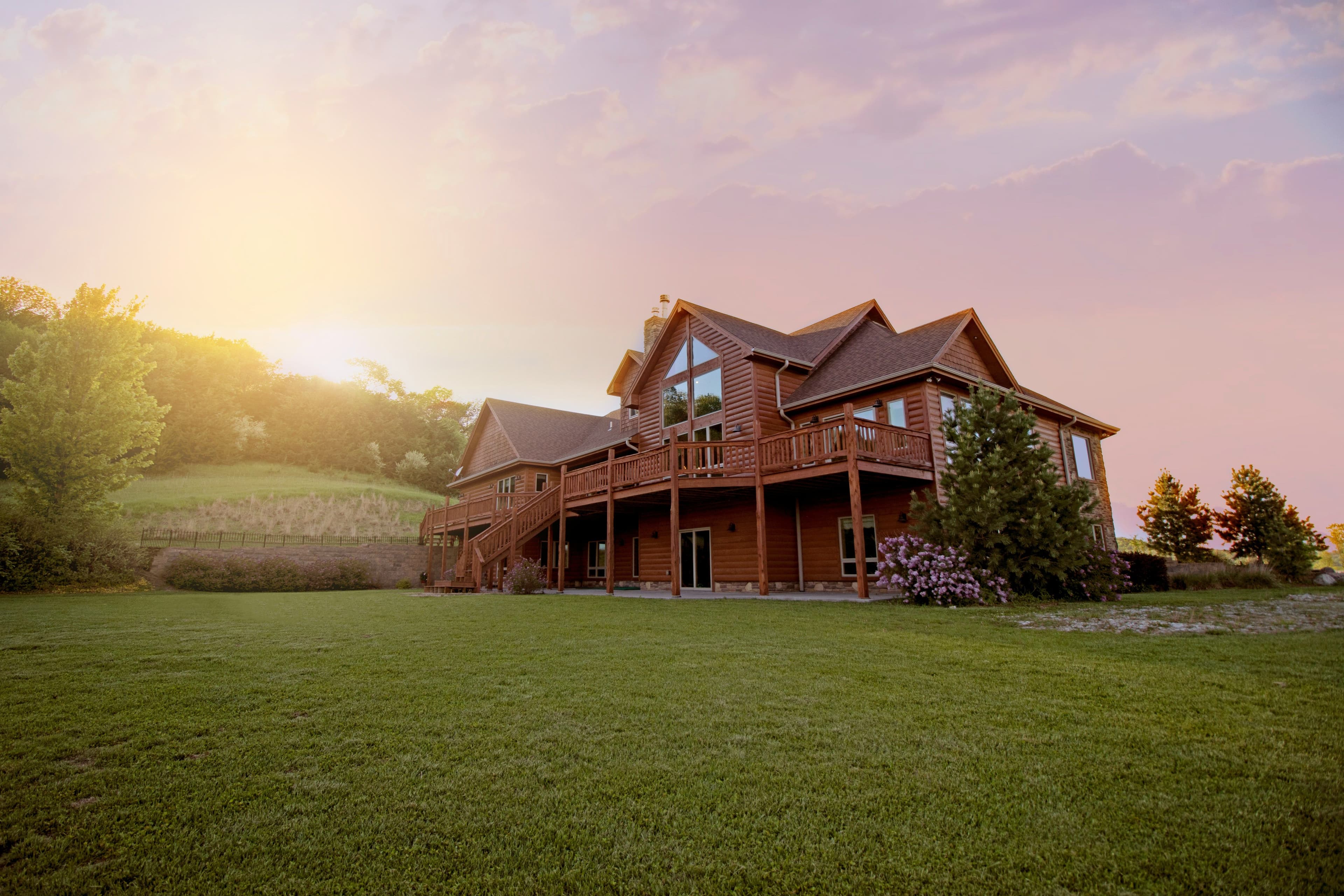 Grande maison en bois avec terrasse surélevée, entourée de pelouses verdoyantes et d'arbres, au coucher du soleil.