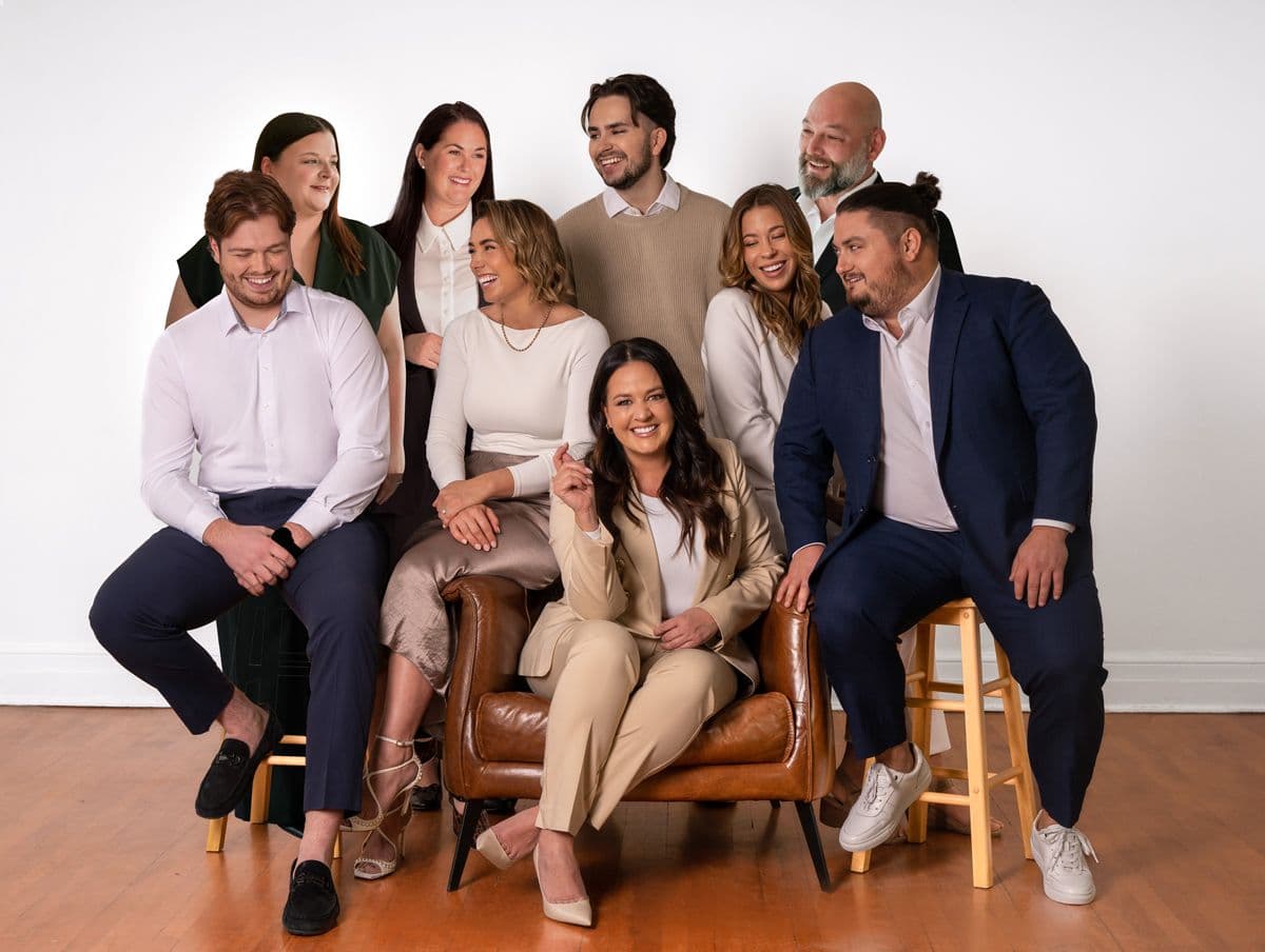 Groupe de neuf personnes souriantes posant dans un studio, habillées de tenues professionnelles dans des tons neutres, sur des chaises et un fauteuil en cuir.