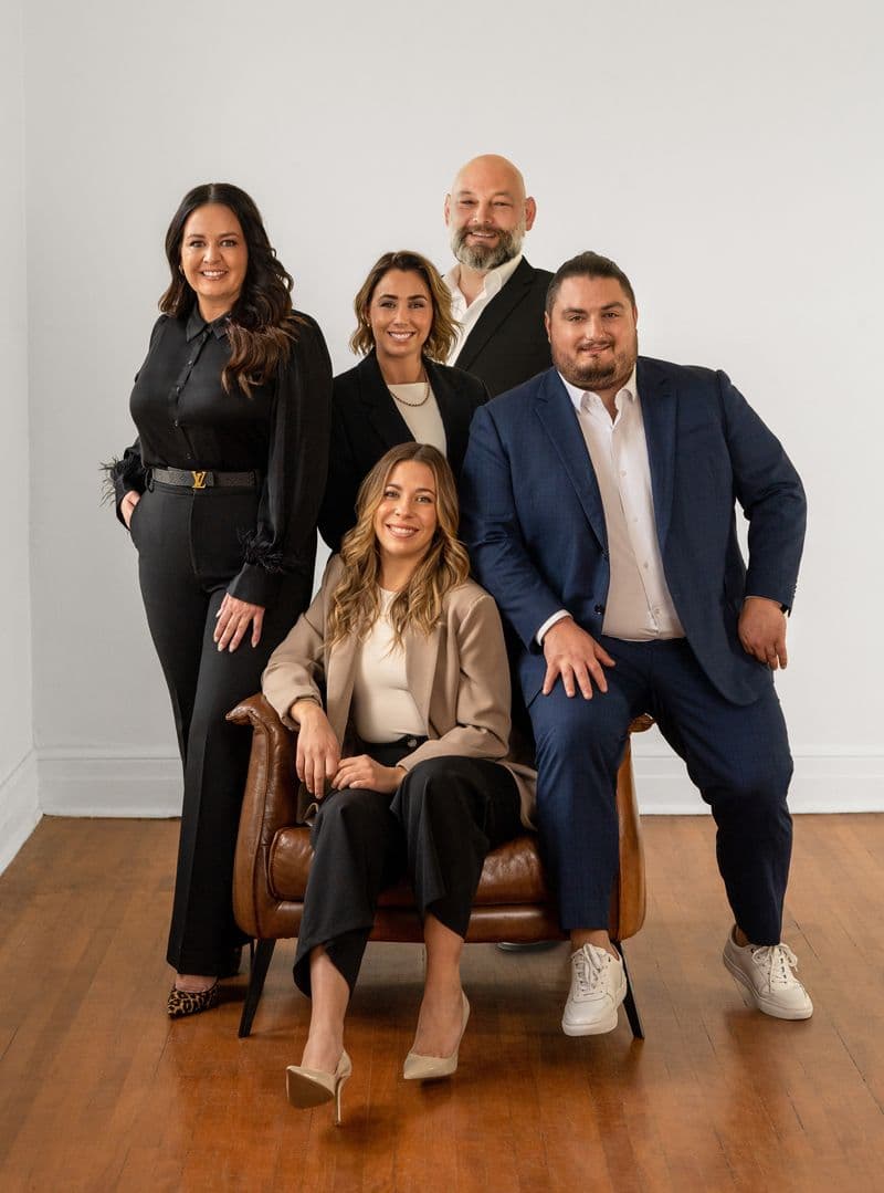 Groupe de quatre professionnels souriants en tenue élégante, posant dans une pièce lumineuse avec un fauteuil en cuir marron
