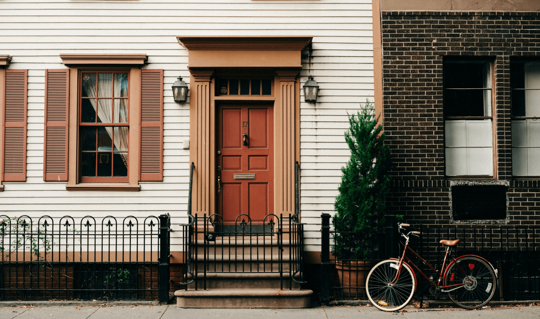 Façade de maison avec une porte rouge entourée de volets assortis, une clôture en fer forgé et un vélo rouge appuyé contre un mur en briques.