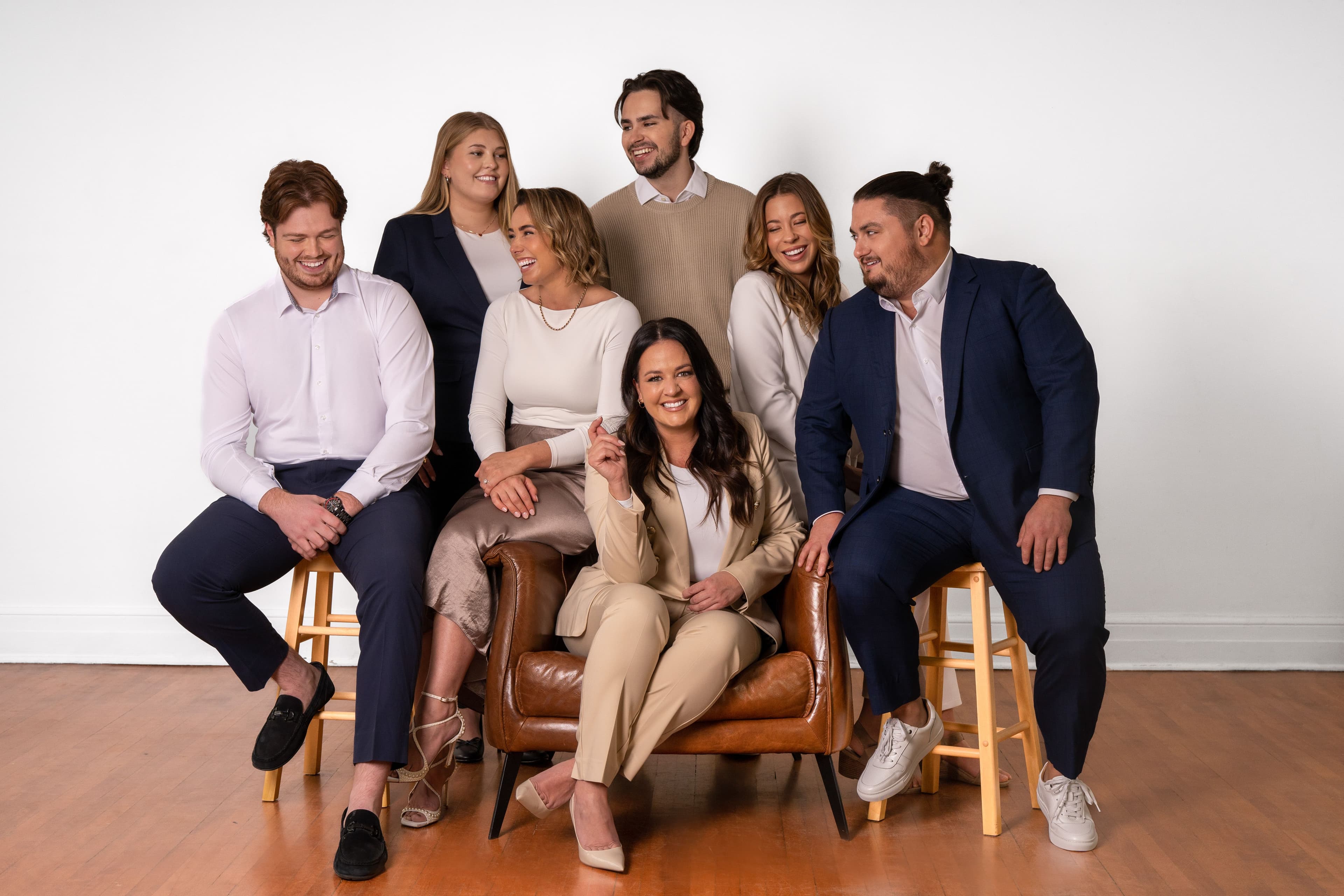 Groupe de sept personnes souriantes posant dans un studio, habillées de tenues professionnelles dans des tons neutres, sur des chaises et un fauteuil en cuir.