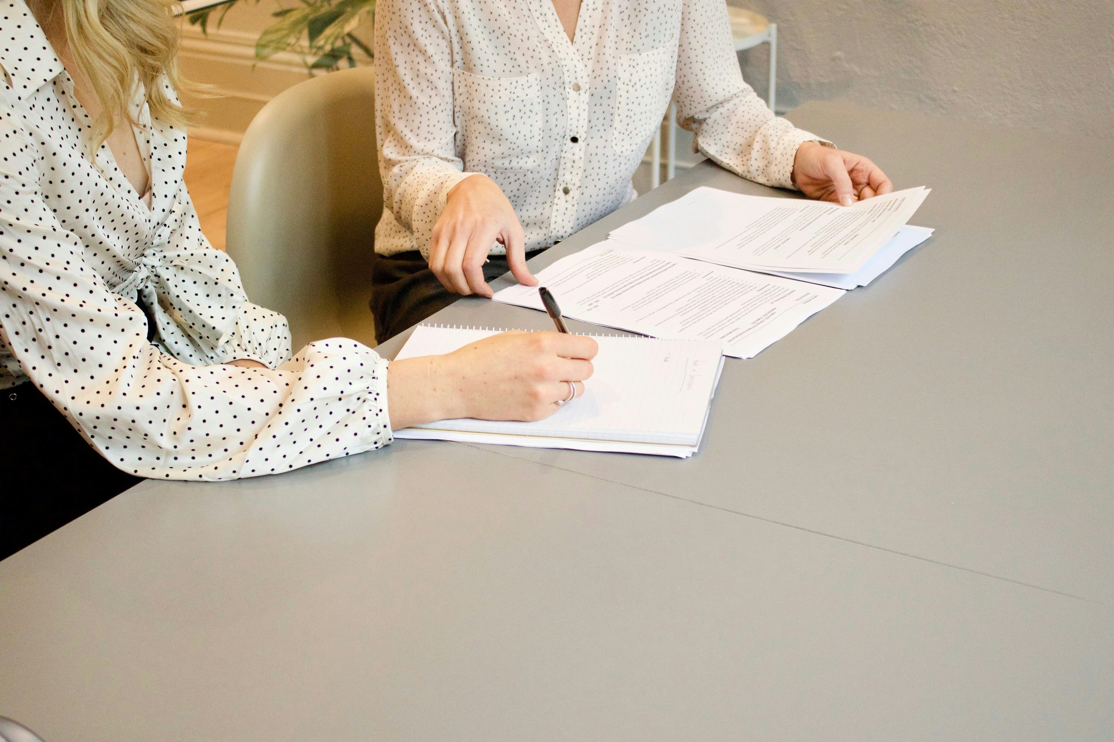 Deux femmes en chemisiers à motifs travaillant sur des documents posés sur une table grise, l'une écrivant avec un stylo.